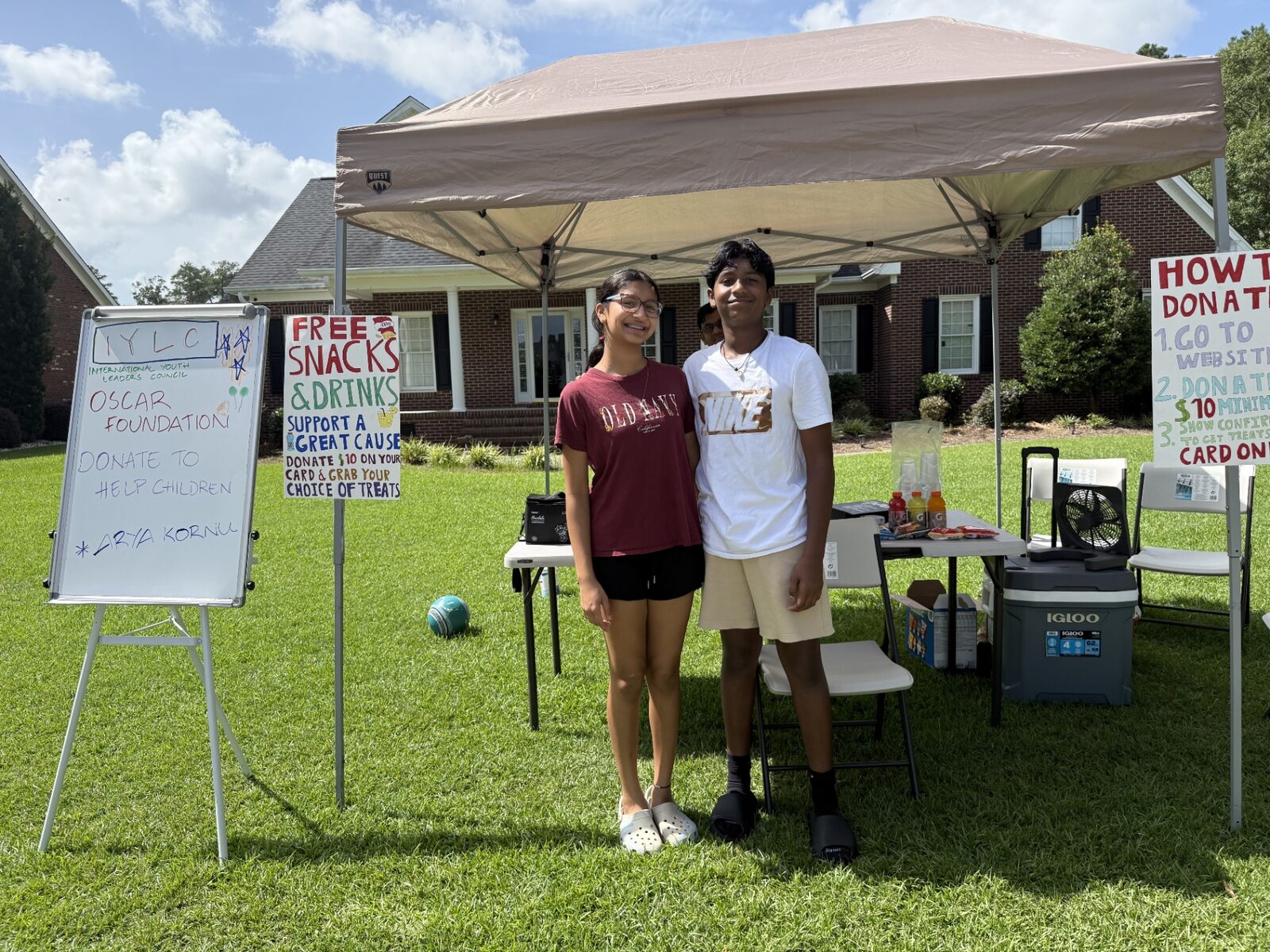 Two students standing under a canopy beside handmade IYLC and OSCAR Foundation signs at a snacks and drinks fundraising stand